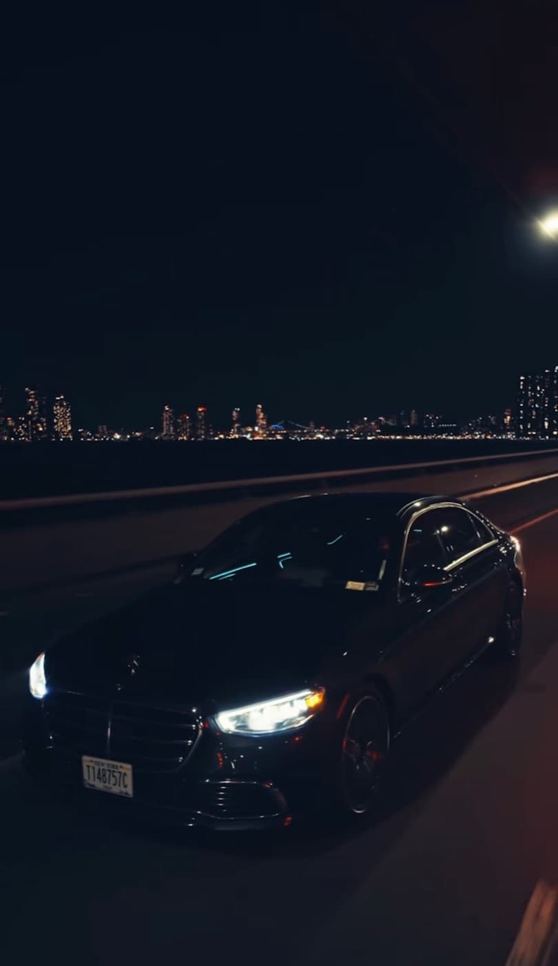 Black Mercedes-Benz S-Class driving at night with the New York City skyline behind